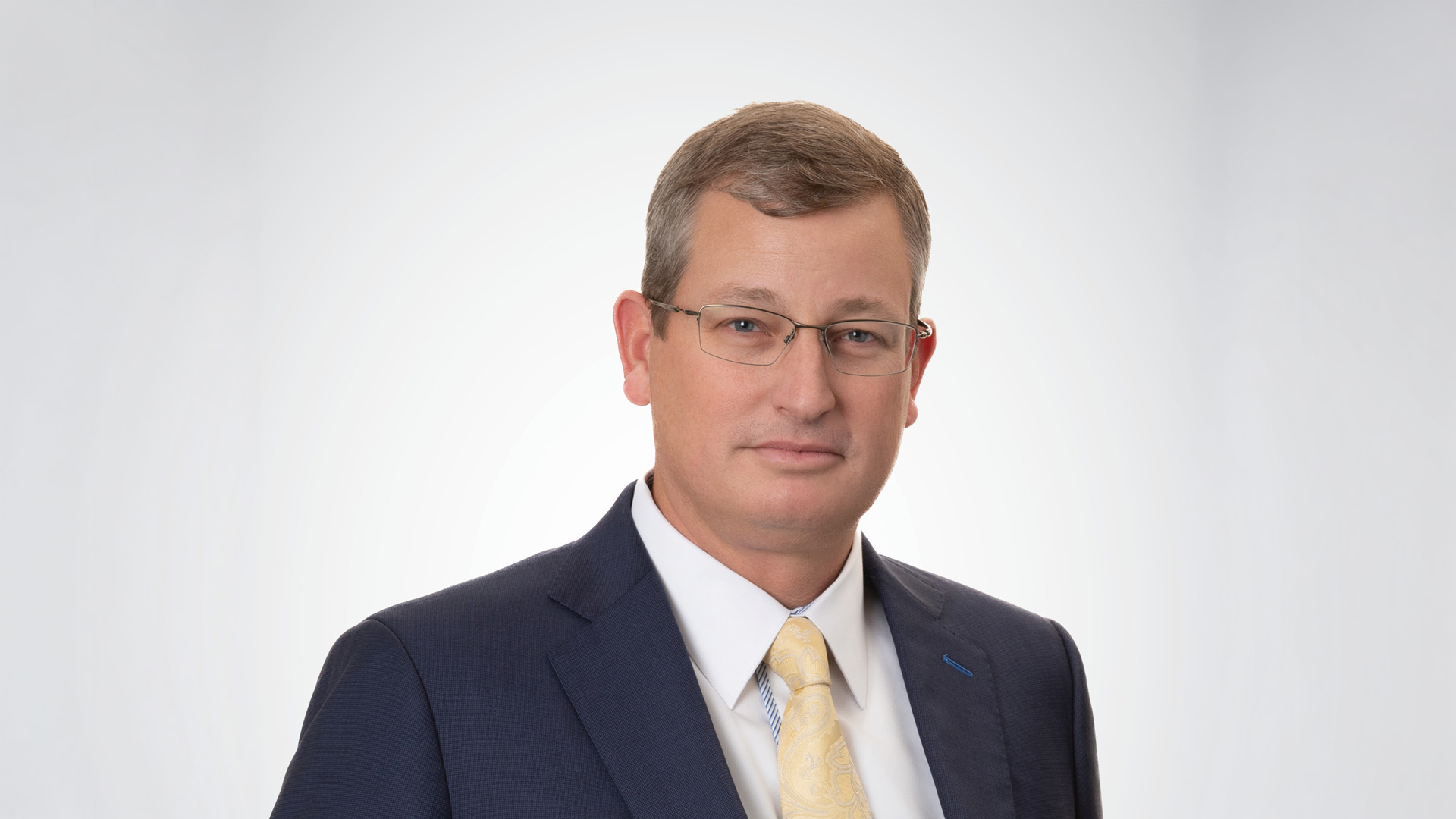 Professional headshot of a man wearing glasses, a navy suit, white shirt, and light yellow tie, facing the camera against a light gray background.